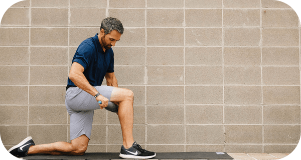 Man using RAD Muscle Flushing Kit | RAD for muscle recovery on thigh while kneeling on a mat against a gray brick wall, promoting wellness and body care.