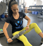 Woman using a gray muscle massage roller on her thigh in a gym setting, demonstrating the RAD Muscle Flushing Kit | RAD for recovery and wellness tools.