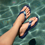Feet wearing navy blue flip-flops with turquoise sea turtle embellishments floating above clear water, showcasing Theresa | Oka-B sandals in a relaxed, spa-inspired setting.
